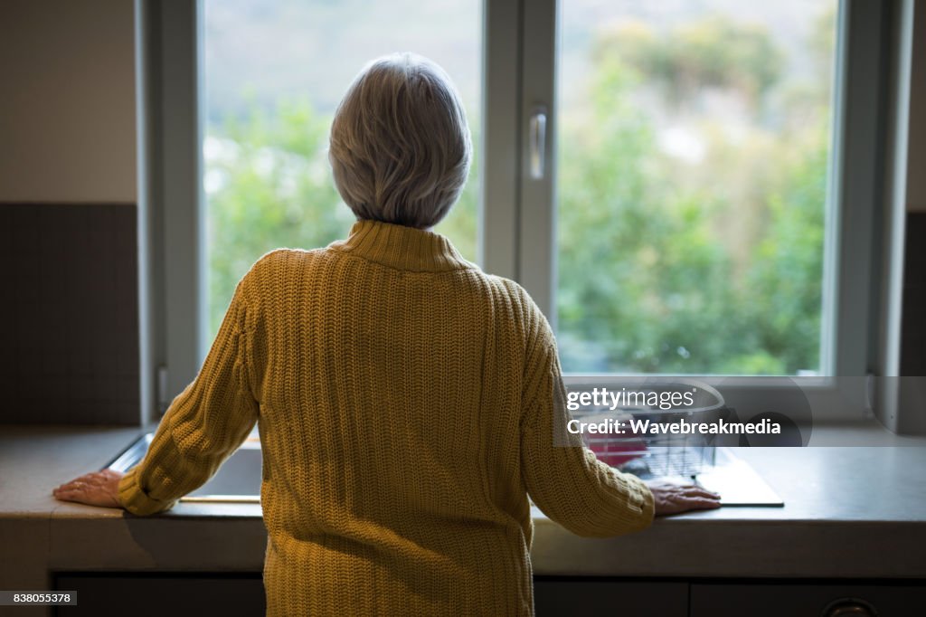 Mujer Senior de pie cerca del fregadero de la cocina y mirando por la ventana