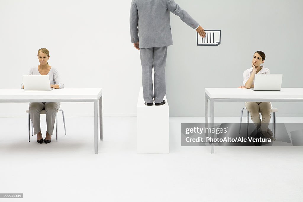 Professional women working with laptops, man standing between them, handing document to one