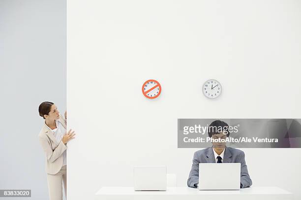 female office worker peeking around corner at clocks on wall, colleague looking down at laptop - looking around stock pictures, royalty-free photos & images