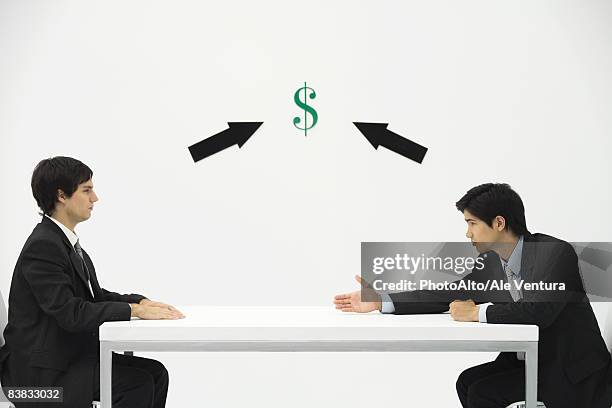 two businessmen face to face, one reaching to offer his hand, dollar sign and arrows between them - uncompromising photos et images de collection