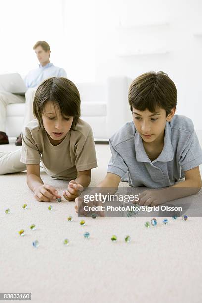 boys lying on the floor playing with marbles, father sitting on sofa with laptop in background - boy sitting on floor stock pictures, royalty-free photos & images