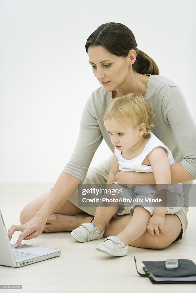 Professional woman sitting on the ground, using laptop, holding toddler on lap