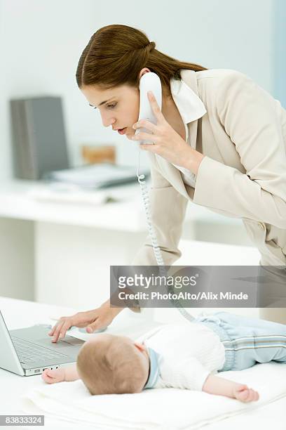 professional woman using phone and laptop computer, baby lying on desk beside her - vornüber beugen stock-fotos und bilder