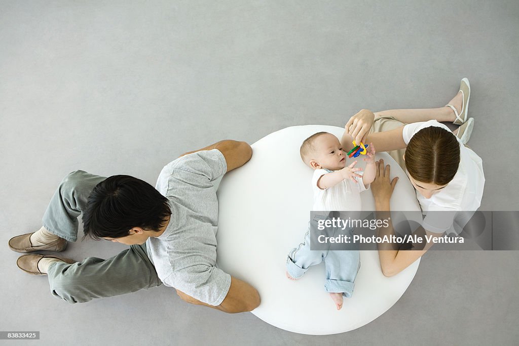 Infant lying on ottoman, mother holding keys over him, father sitting with back turned, overhead view