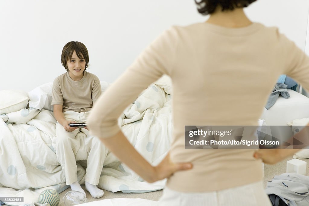Boy playing handheld video game in messy bedroom, mother standing in foreground with hands on hips
