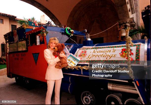 Portrait of American actress Marla Maples hugs a teddy bear as she poses beside a vehicle, decorated as a Christmas train with a banner that reads...