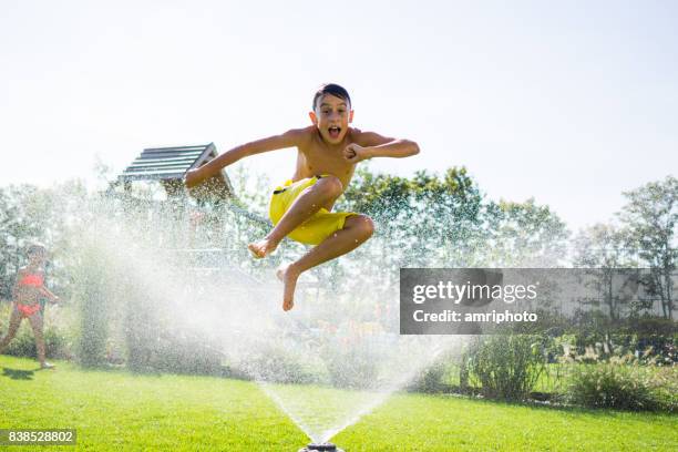 boy jumping over garden sprinkler on sunny summer day at home - jumping sprinkler stock pictures, royalty-free photos & images
