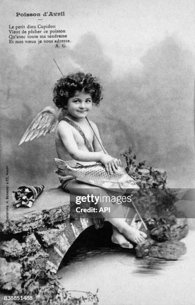 Carte postale illustrée par la photographie de Cupidon assis sur un pont et un poisson pour le 1er avril, en France.