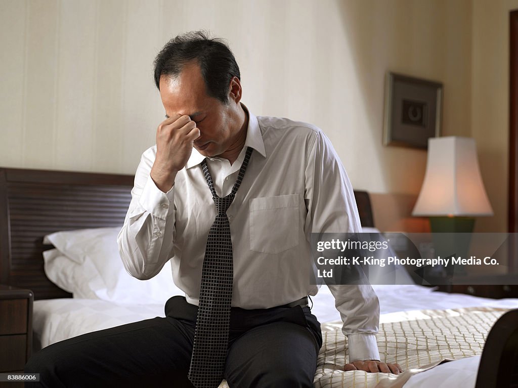 Businessman sitting on side of bed, looking tired.