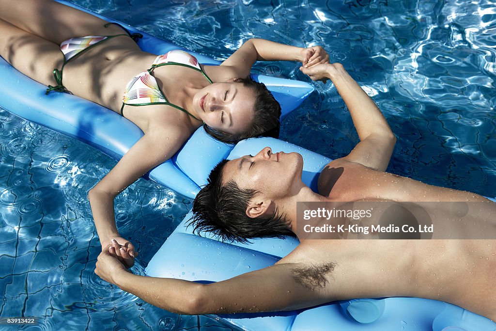 Couple floating on rafts in pool, holding hands.