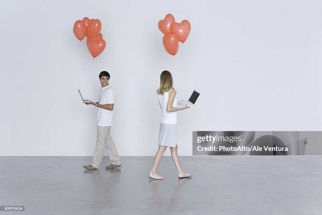 Man and woman walking past each other, both carrying laptop computers and heart balloons