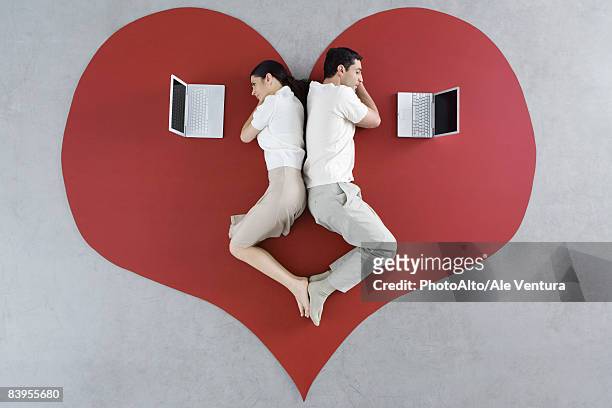 man and woman lying back to back on large heart, both looking at laptop computers - relation à distance photos et images de collection
