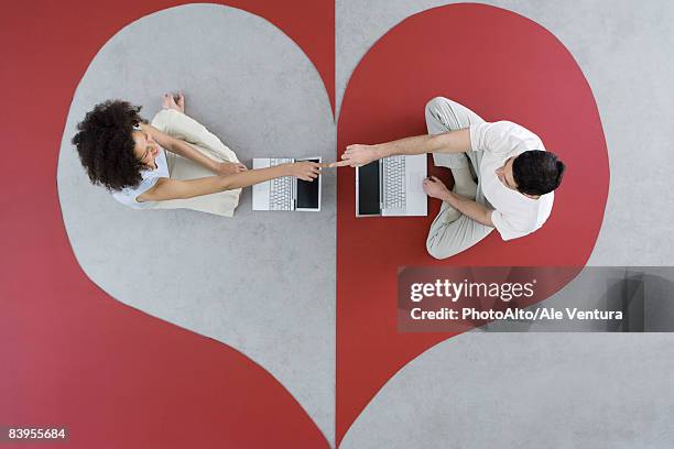 couple sitting face to face with laptop computers on heart shape, touching fingers, overhead view - tinder stock-fotos und bilder