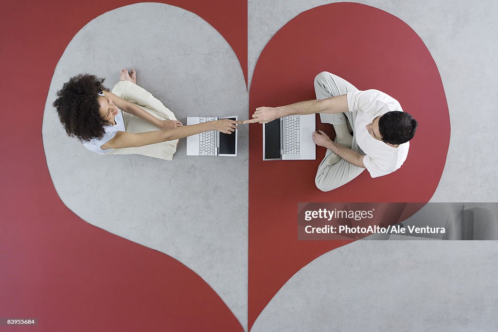 Couple sitting face to face with laptop computers on heart shape, touching fingers, overhead view