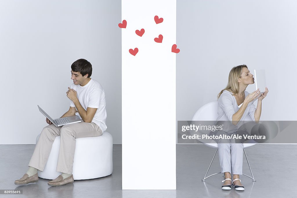 Woman kissing laptop computer, man looking at his own laptop and smiling, hearts in the air between them