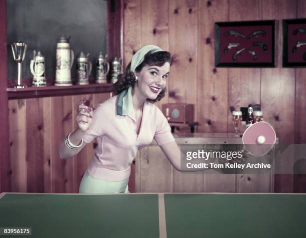 Woman smiles as she returns a shot in ping pong while playing in a wood-panelled, beer-stein decorated game room, ca.1950s. United States.