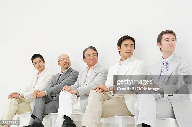 businessmen sitting on chairs in row against wall, looking right, legs crossed - legs crossed at knee stock pictures, royalty-free photos & images