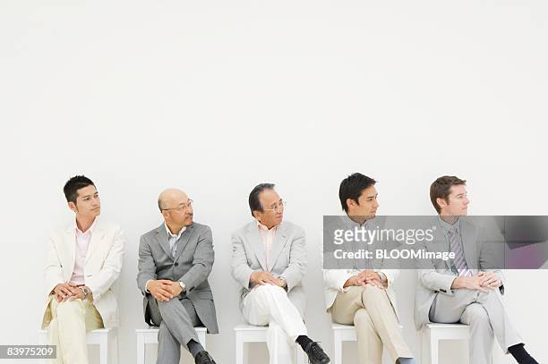 businessmen sitting on chairs in row against wall, looking right, legs crossed - legs crossed at knee stock pictures, royalty-free photos & images
