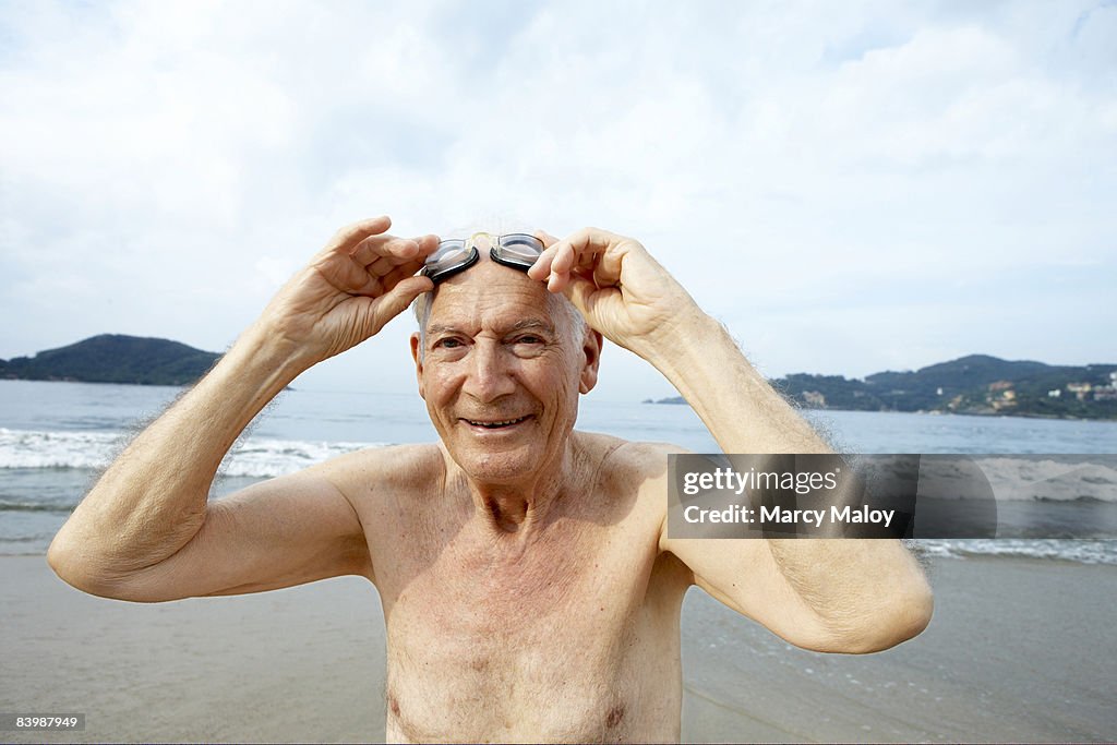 Elderly man on beach holding goggles above eyes.