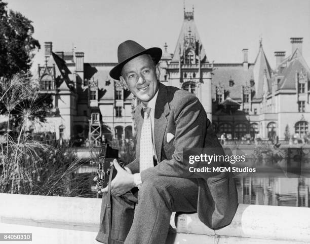 British actor Alec Guinness holds a Rolleiflex camera while seated near a pond in front of the Biltmore House during the shooting of the film 'The...