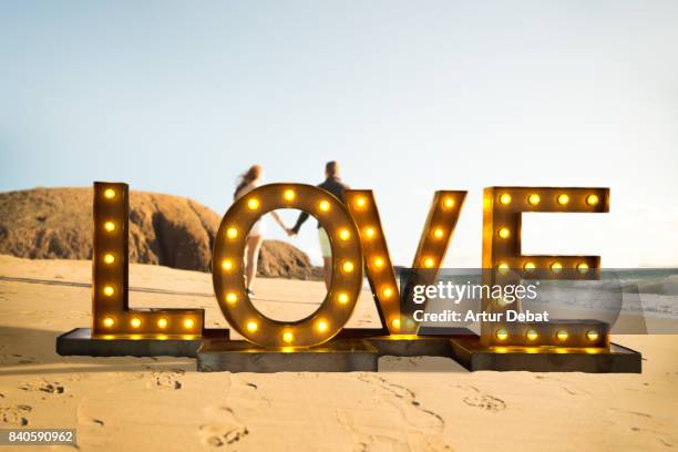 creative creation of a love letters illuminated with light bulbs in front a happy couple in love walking in the canary islands beach with nice light during travel vacations in spain. - light bulb letters stock pictures, royalty-free photos & images