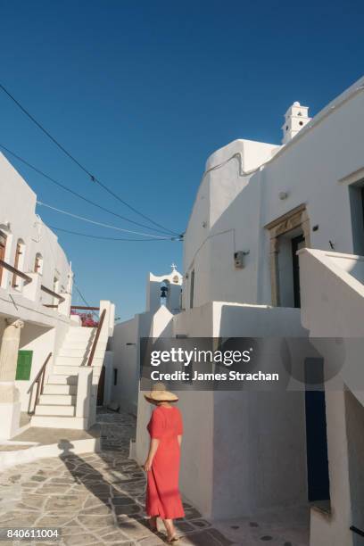 woman in red dress and straw hat walks though traditional white greek houses (by church with crooked cross) in evening light casting a long shadow, kastro village, sifnos, cyclades islands, greece - sifnos stock-fotos und bilder