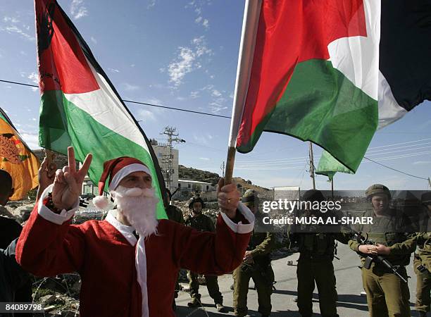 Palestinian protestor dressed as Santa Claus or Father Christmas holds a Palestinian flag and flashes a "V" for victory sign as Israeli soldiers look...