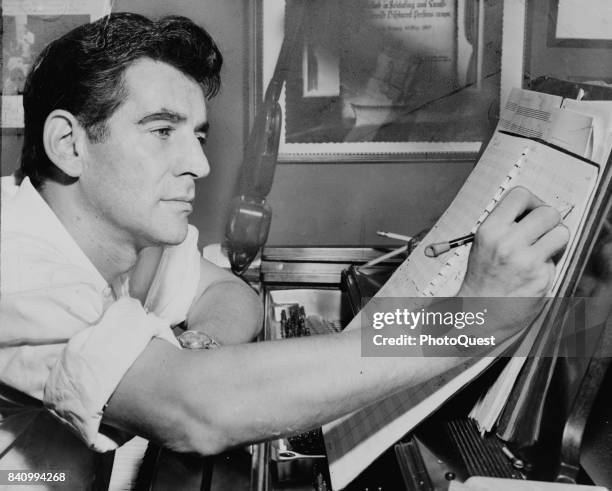 View of American composer Leonard Bernstein as he sits at a piano and makes notations to a musical score, New York, New York, 1955.