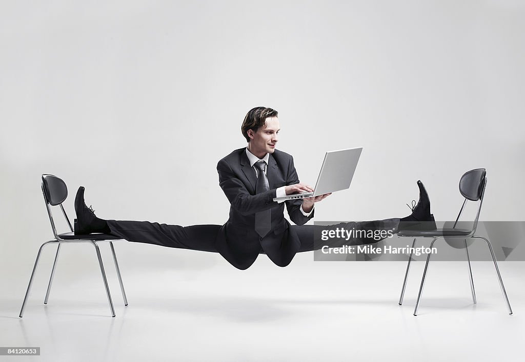 Businessman balancing to two chairs holding laptop