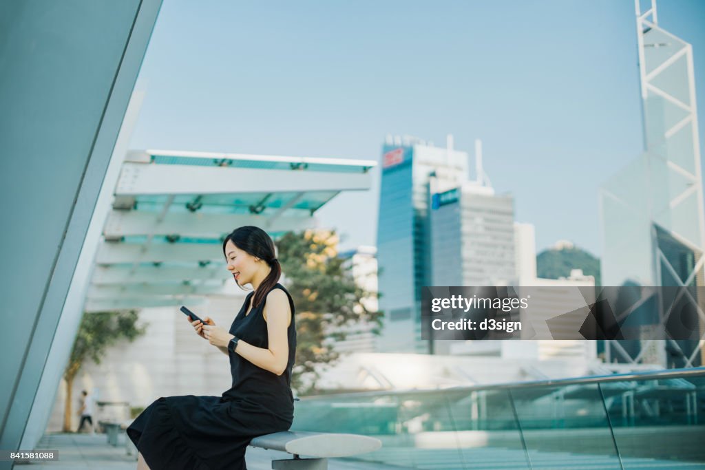 Young woman using smartphone on urban balcony, with highrise corporate buildings behind as background