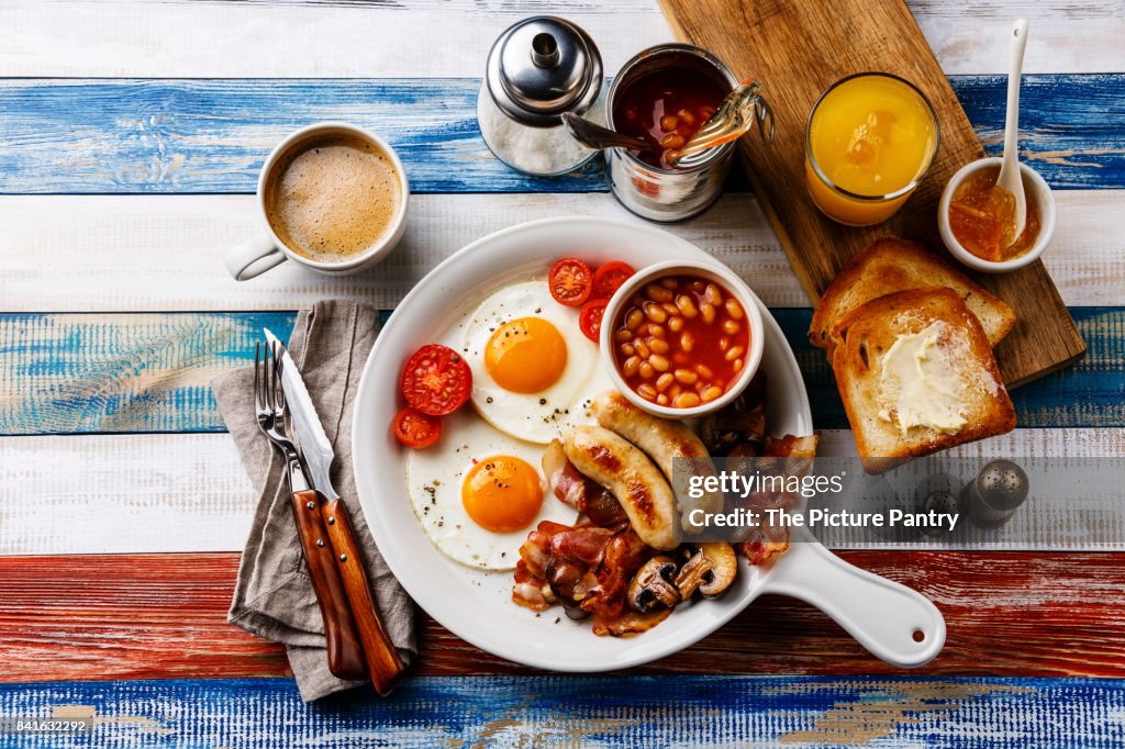Full English breakfast in white pan with fried eggs, sausages, bacon, beans, toasts, orange fresh and coffee on wooden background