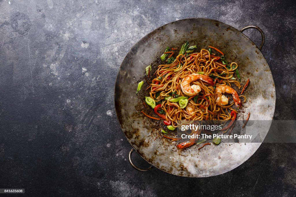 Udon noodles stir-fried with Tiger shrimps and vegetable in wok cooking pan on dark background copy space