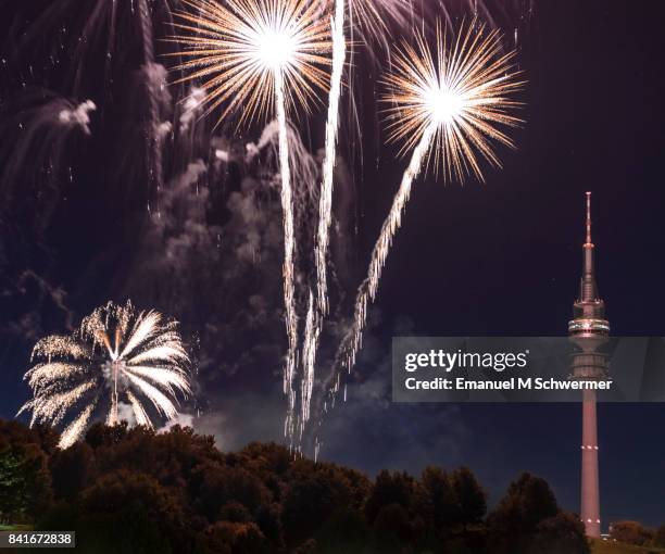 spectacular, majestic fireworks display with colorful starburst explosives exploding in the night sky, rockets shot up bursting, glowing, sparkling and streaking lights during summer festival with munich’s . tv tower in background illuminated - olympiaturm stock-fotos und bilder