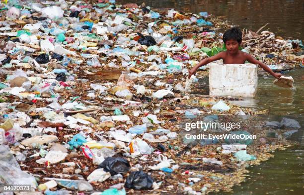 Young poor boy rowing in his boat made of styropor is collecting cans and plastic bottles out of the garbage swimming in the Siem Reap River near the...