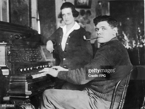 American baseball player Babe Ruth smokes a pipe as he plays a piano while his wife, Helen Ruth stands next to him, 1910s.
