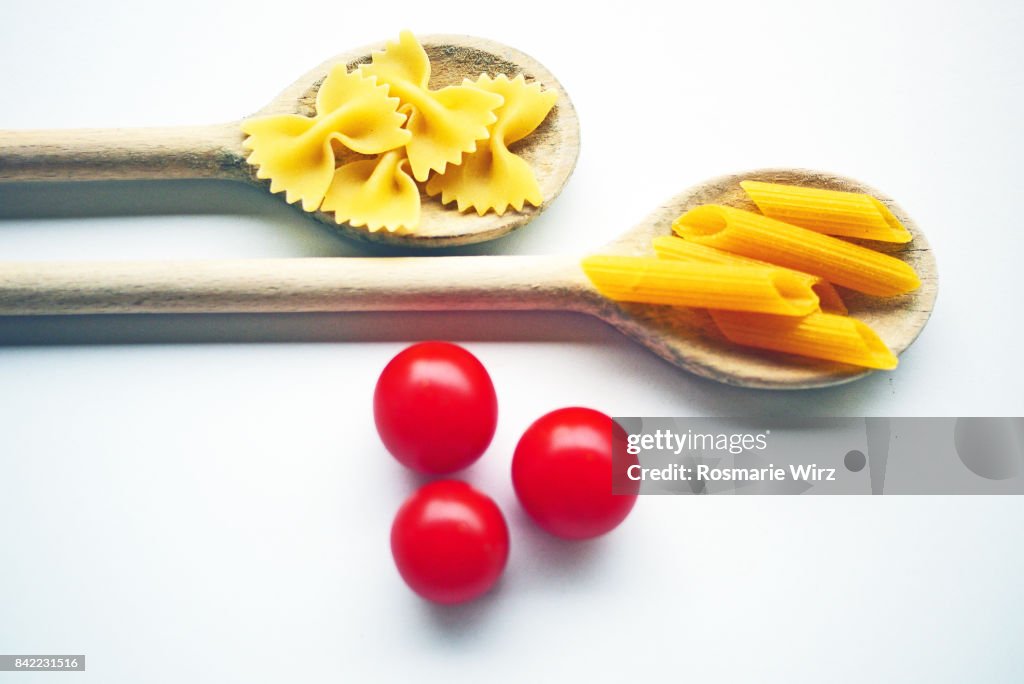 Shapes of Italian raw pasta on wooden spoons, cherry tomatoes, on white.