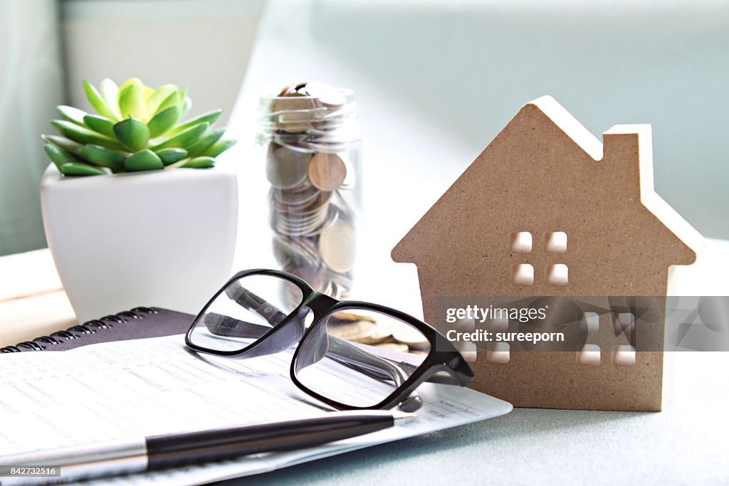 Wood house model, coins, eyeglasses and saving account book or financial statement on office desk table