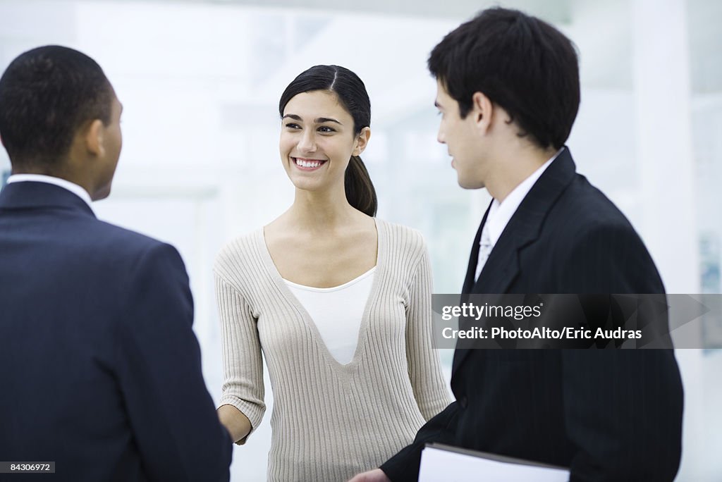 Professional woman standing with two businessman, shaking one man's hand, smiling