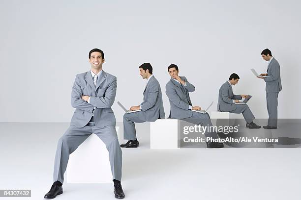 businessman smiling at camera with arms folded while his clones use laptops in background - multiplication photos et images de collection