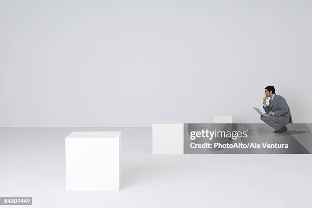 businessman squatting using laptop, looking at empty pedestals - mid distance stockfoto's en -beelden