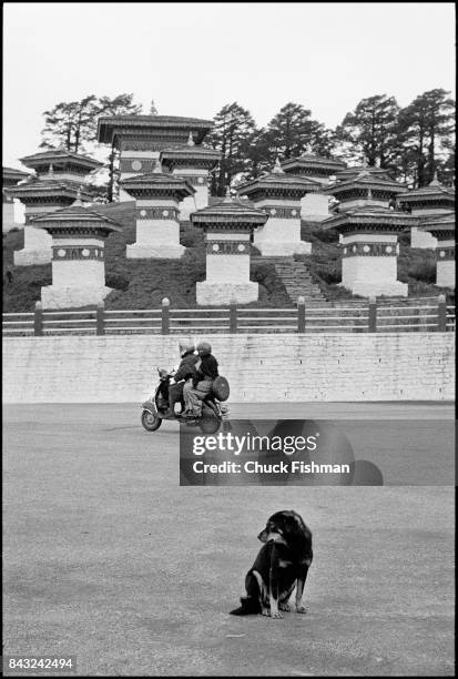 As a dog sits on the road in the foreground, a couple ride a moped past the Druk Wangyal Chortens in the Dochula Pass, Bhutan, November 2004. The...