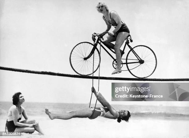 Nouvel exercice inventé par les Beach Girls: une jeune femme fait du vélo sur une corde tendue tandis qu'une deuxième fait contrepoids pour...