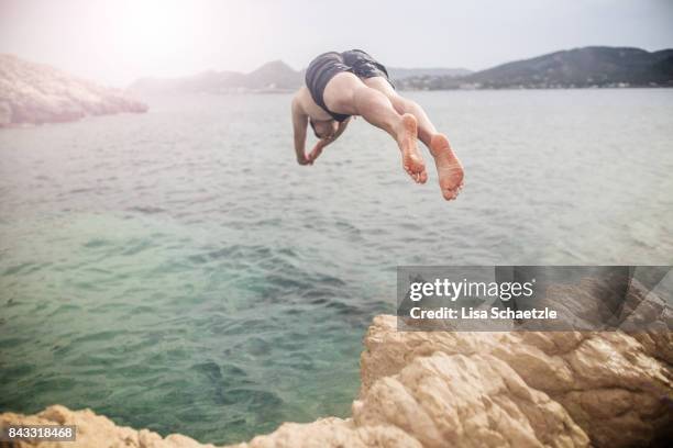 young man jumping off cliff into water - klippenspringen stock-fotos und bilder