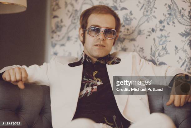 British singer and musician Elton John, wearing a white suit, black shirt with flower motif and multicolored sunglasses, London, November 1973.