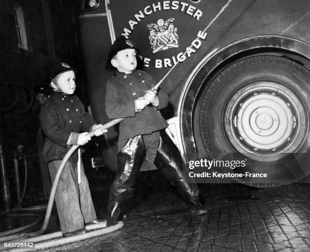 Dans une caserne de pompier, deux jeunes garçons vêtus de l'uniforme tirent sur le tuyau de la lance d'incendie, à Manchester, Royaume-Uni en 1961.