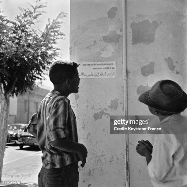 Regroupement de populations civiles en Oranie, deux jeunes hommes lisent une affichette placardée sur un mur où il est écrit en français et en arabe...
