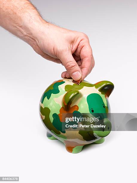 hand putting coin into camouflage piggy bank - saving for a rainy day englische redewendung stock-fotos und bilder