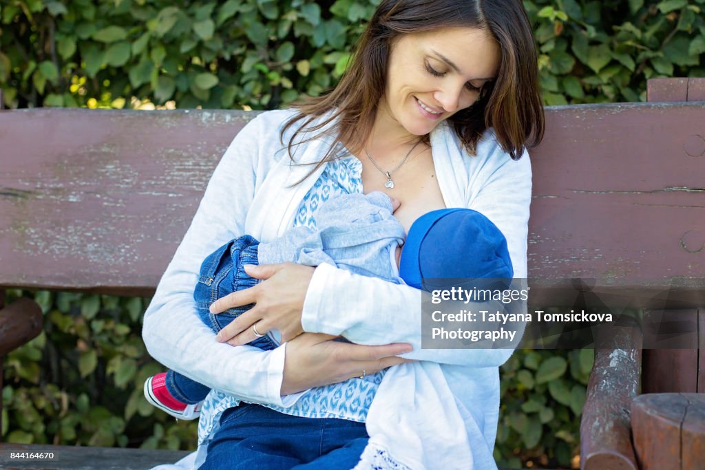 Young beautiful mother sits on bench, breastfeeding her newborn baby boy in the park