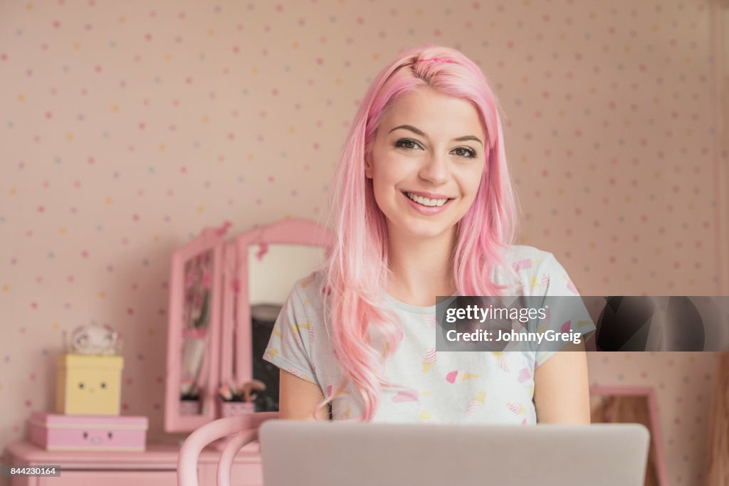 Cheerful young woman with pink hair using laptop in bedroom