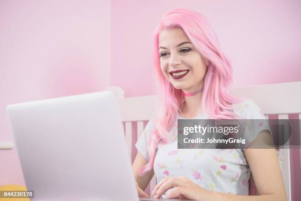 mujer joven con el pelo teñido de rosa usando laptop y sonriendo hacia el monitor - dyed red hair fotografías e imágenes de stock